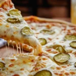 A hand holding a cheesy slice of pickle pie pizza with melted mozzarella and dill pickles, showcasing a perfect cheese pull.